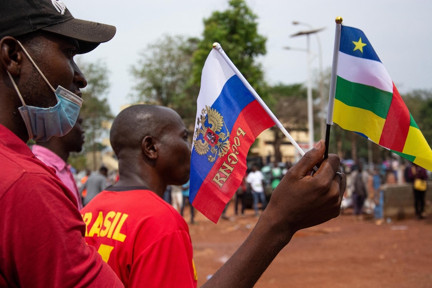 A man with a face mask pulled under his nose and mouth waves Russian and Central African Republic flags