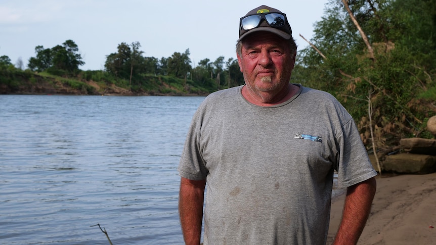 A man wearing a grey t-shirt and hat looks at the camera next to a river