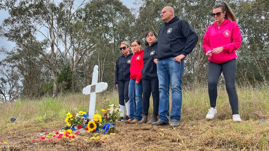 A group of people gather around a crucifix placed in the ground, which is surrounded by flowers.