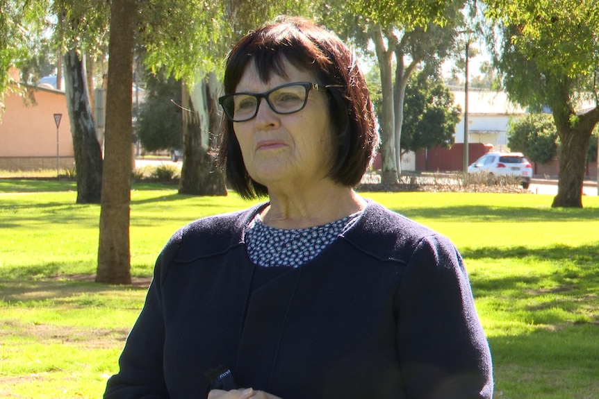 A dark haired woman, with a dark top on standing in a park on a sunny day