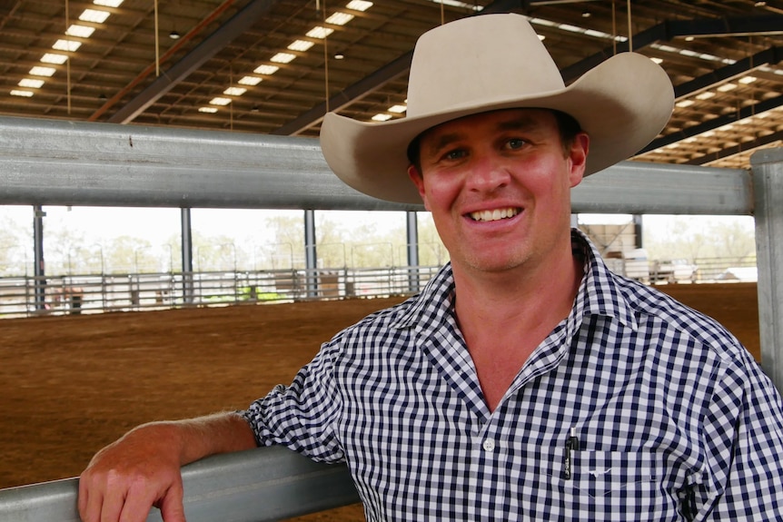 A man in a check shirt wearing a hat leaning against a fence