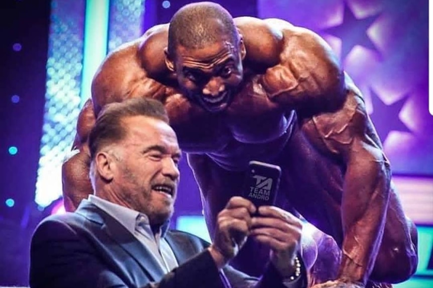 Schwarzenegger stands at the edge of the stage, showing a grinning McMillan something on his phone.