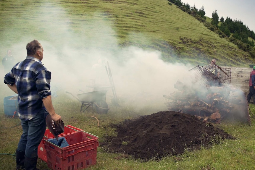 A man watches on as a large fire is built in a paddock.