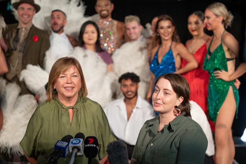 Two women in front of microphones with costumed stage performers in the background