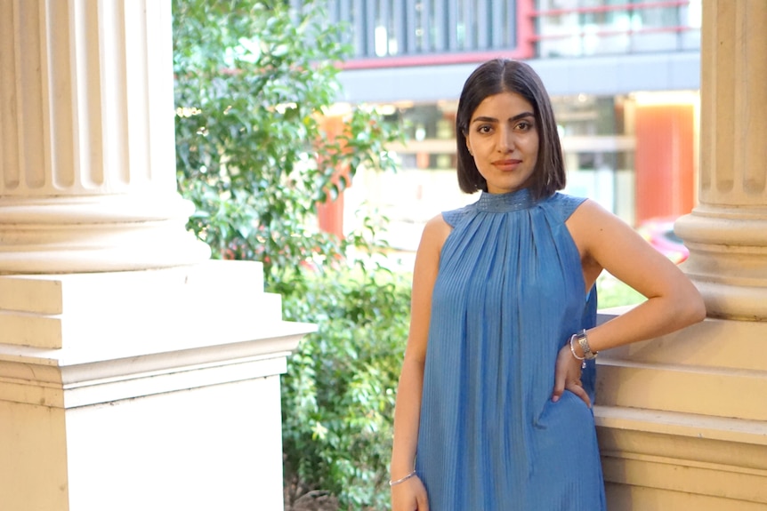 A young Afghan woman wearing a blue high-necked, sleeveless dress poses for a photo.