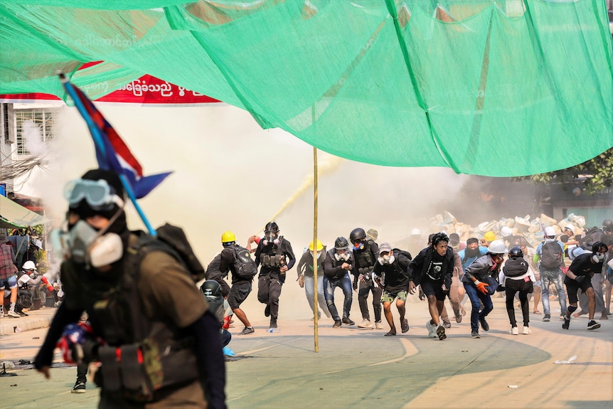 A group of street protesters duck under a green canopy with smoke behind them.