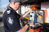 Police officer opening plastic pelican case holding a piece of gear that has a lens under the handle and a keypad on the bottom.