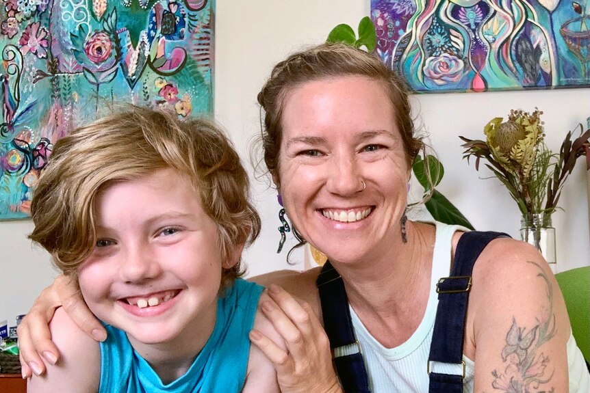 Middle-aged woman with tattoos and a big smile hugs her ten-year-old son with light blue shirt and golden hair in front of art.