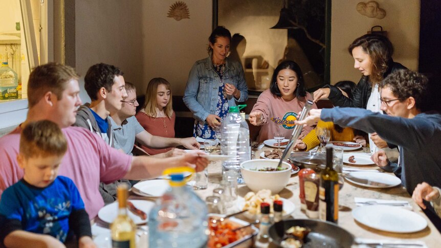 A family group sit and stand around a large table helping themselves to food