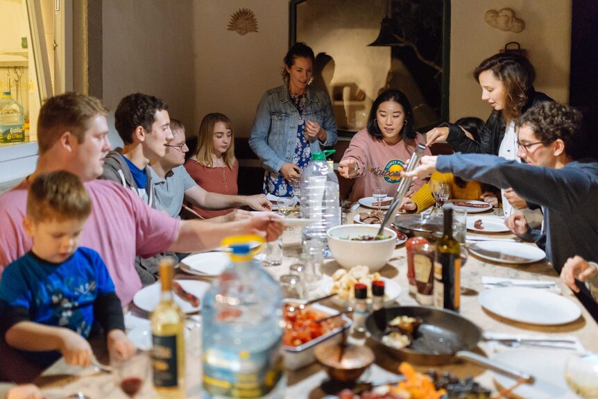 A family group sit and stand around a large table helping themselves to food