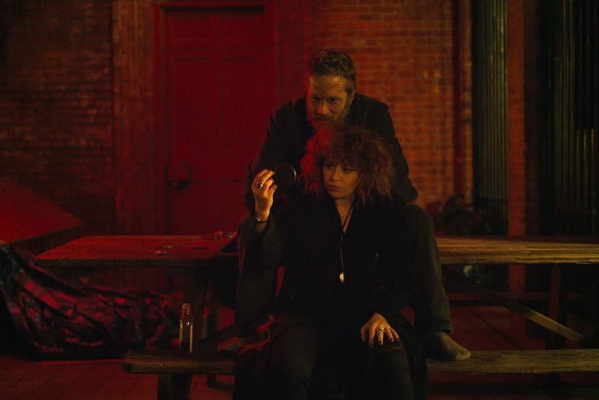 Red-lit nightime scene featuring man and woman sitting at bench outside, brick wall with red door in background.