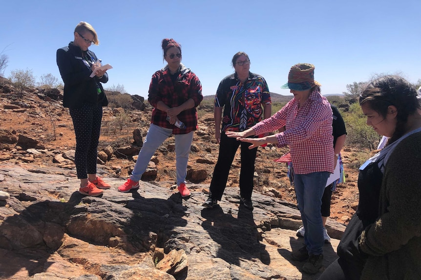 A woman surrounded by five other women, points to a man-made water catchment in rocks.