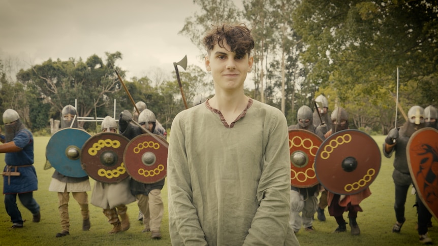 A young man with dark hair in a green tunic standing in front of a line of people in Viking costume charging.