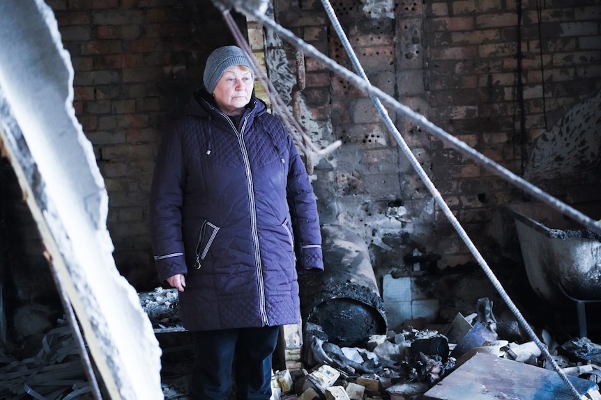 A woman wearing a beanie and warm coat stares at a room with broken furniture and building debris.