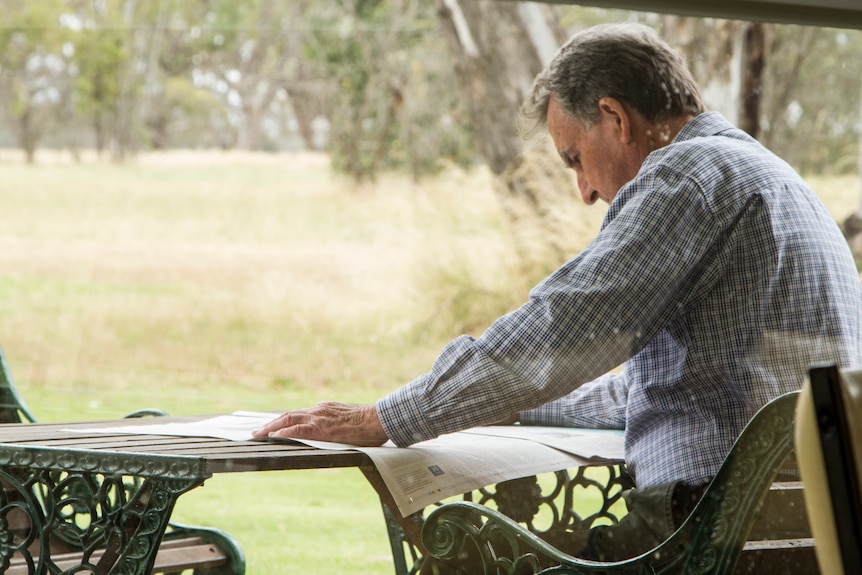 The author reading a newspaper at a bench surrounded by trees.