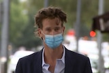 A young man wearing a blue suit jacket, purple shirt and blue face mask walks on a city street