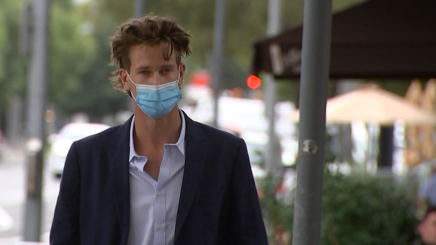 A young man wearing a blue suit jacket, purple shirt and blue face mask walks on a city street