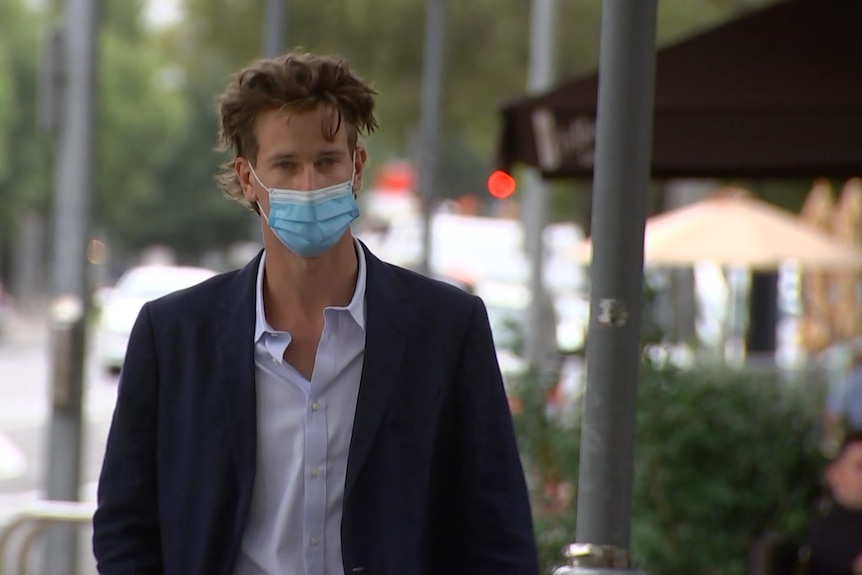 A young man wearing a blue suit jacket, purple shirt and blue face mask walks on a city street