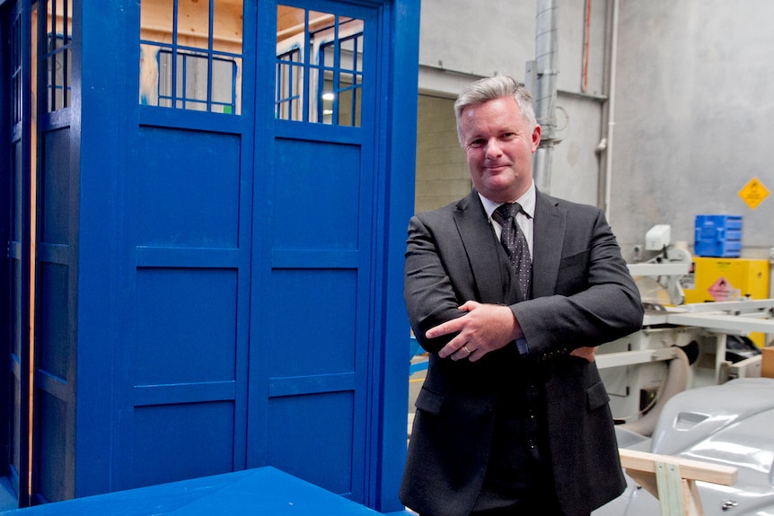 A school principal, dressed in a suit and tie, stands in front of a giant blue box