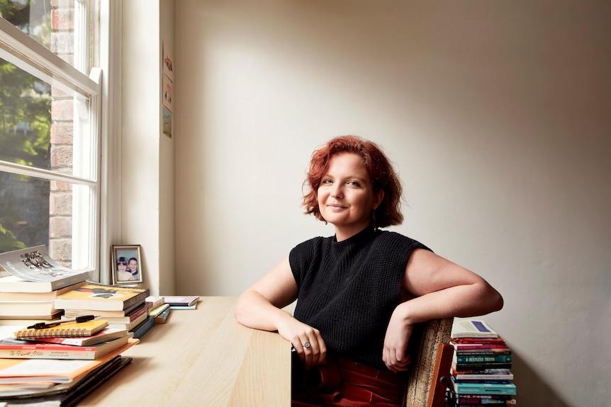 Madeleine Gary con el pelo rojo corto y un top negro sentada en un escritorio con libros delante y detrás