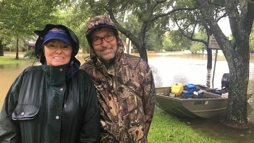 Gloria Thompson and Doug Warwick stand in front of a boat, surrounded by floodwaters.