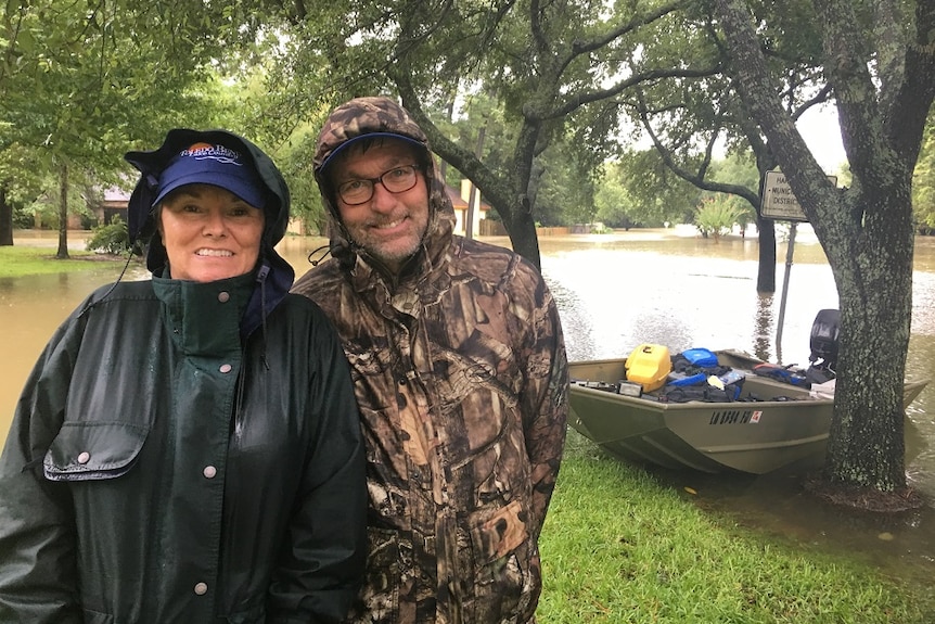 Gloria Thompson and Doug Warwick stand in front of a boat, surrounded by floodwaters.