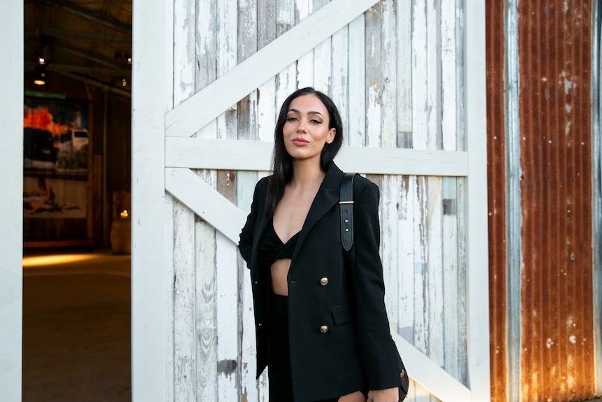 A young woman with dark long hair dressed in black stands in front of a rustic shelter.