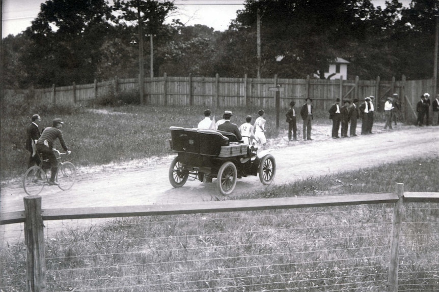A black and white photo of two men running down a dirt road while followed by an old car