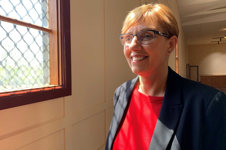 A smiling woman wearing glasses stands near a window.