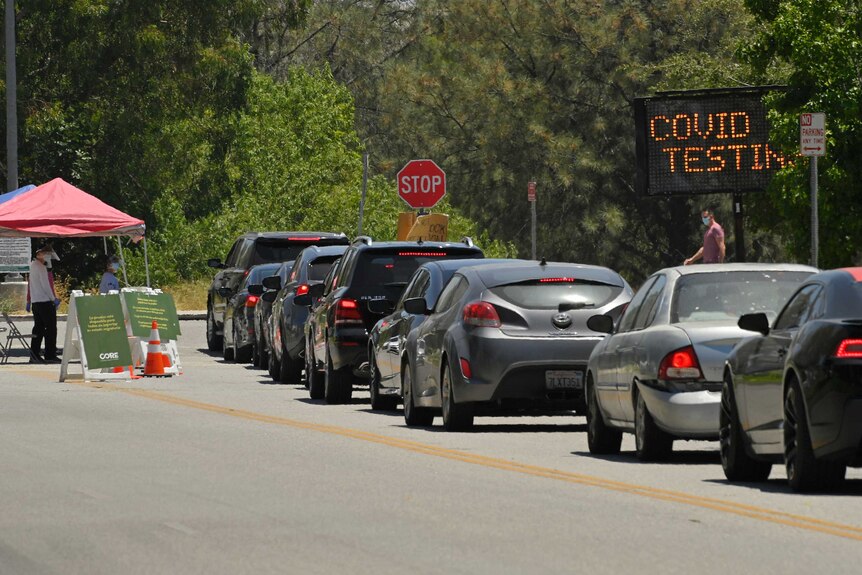 A line of cars queues up at a COVID-19 testing station with masked attendants at booth in greenery.