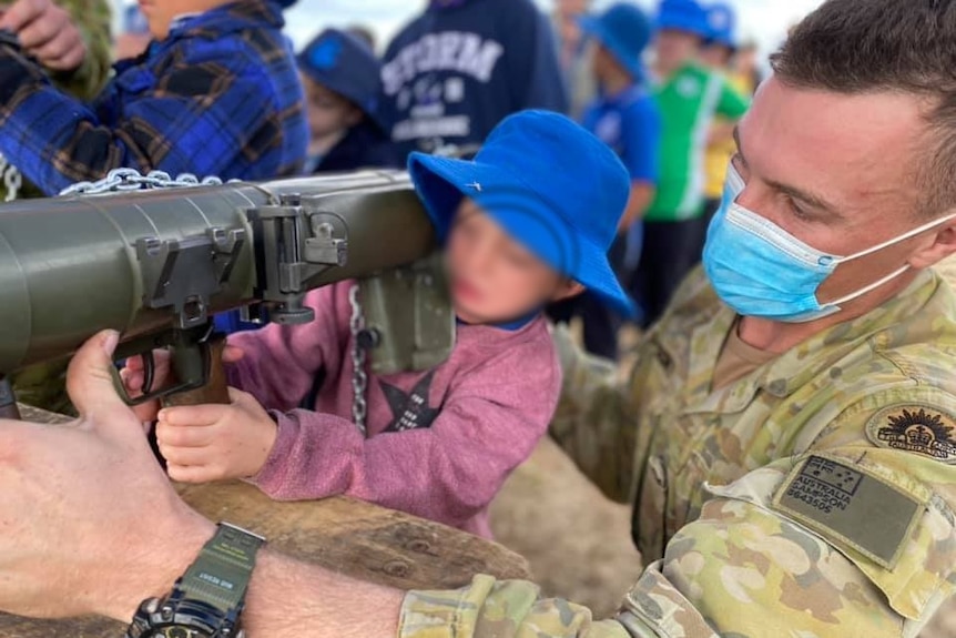 A soldier in fatigues and a mask demonstrates weaponry to a young child wearing a maroon jumper