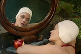 Marion Cotillard in maroon bathers rests her head on the steps of a riverbank and looks longingly at her reflection in a mirror.