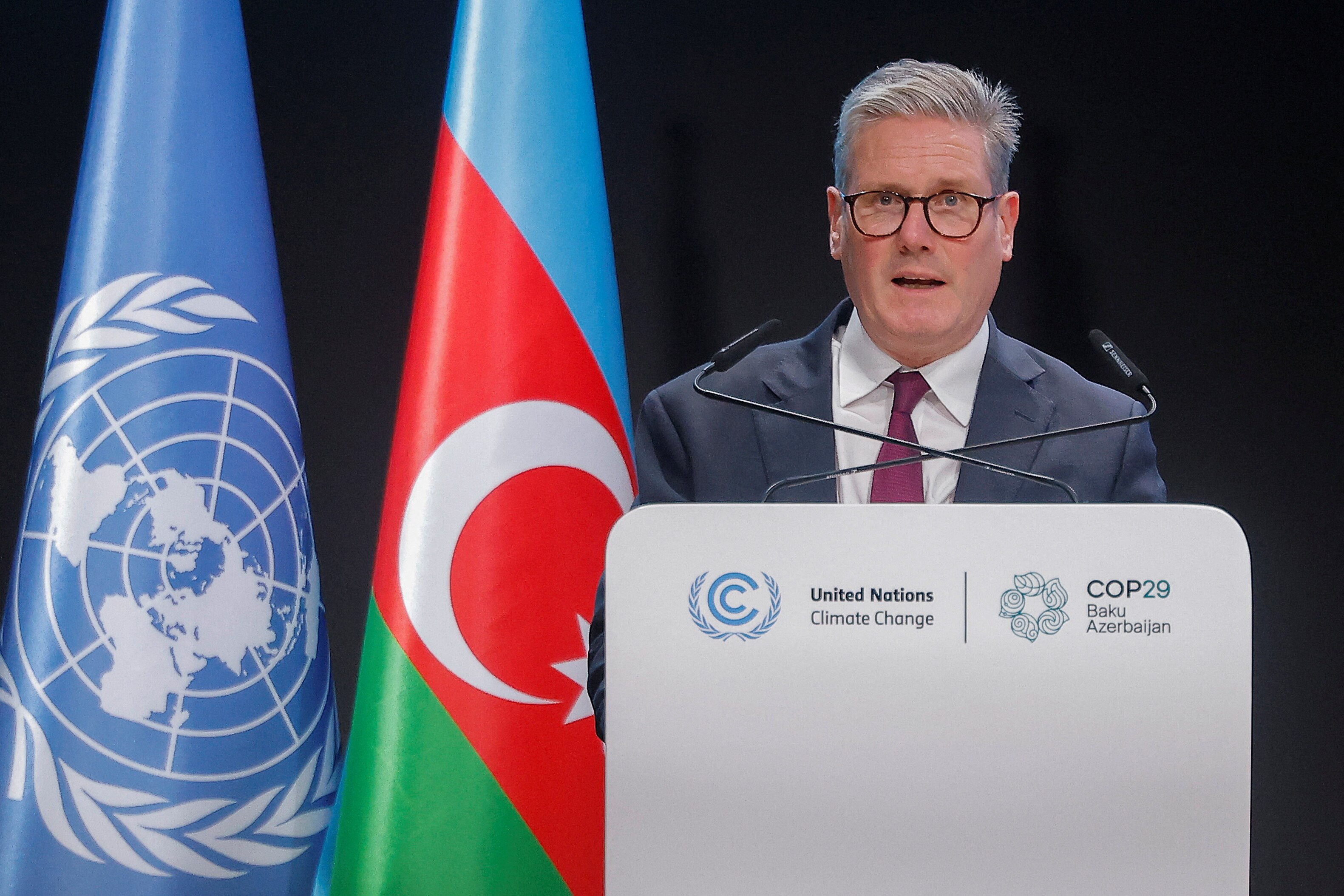 man gives a speech wearing suit with flags in background