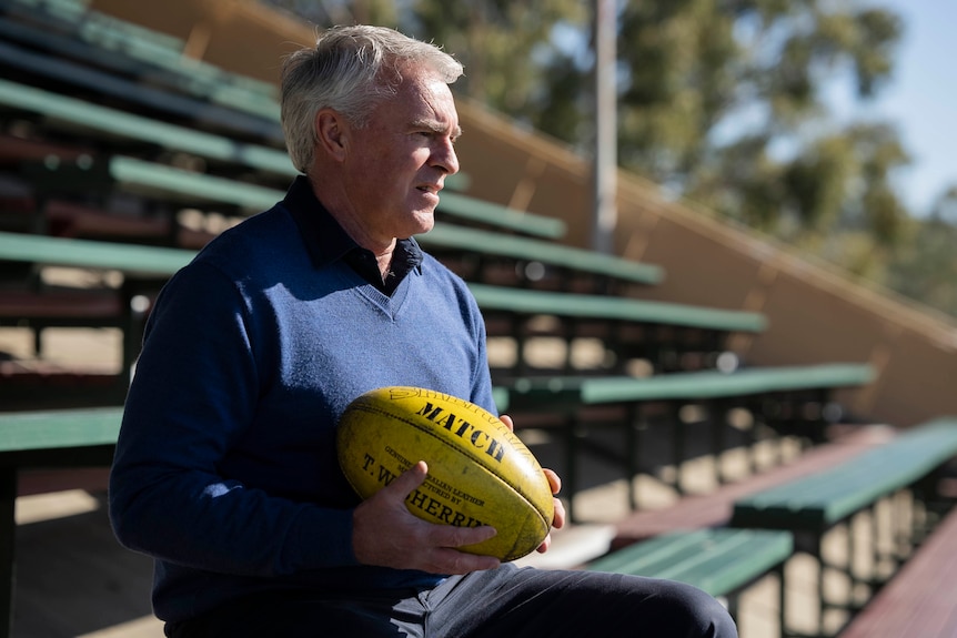 A man with grey hair sits in a sports stand holding and AFL ball.