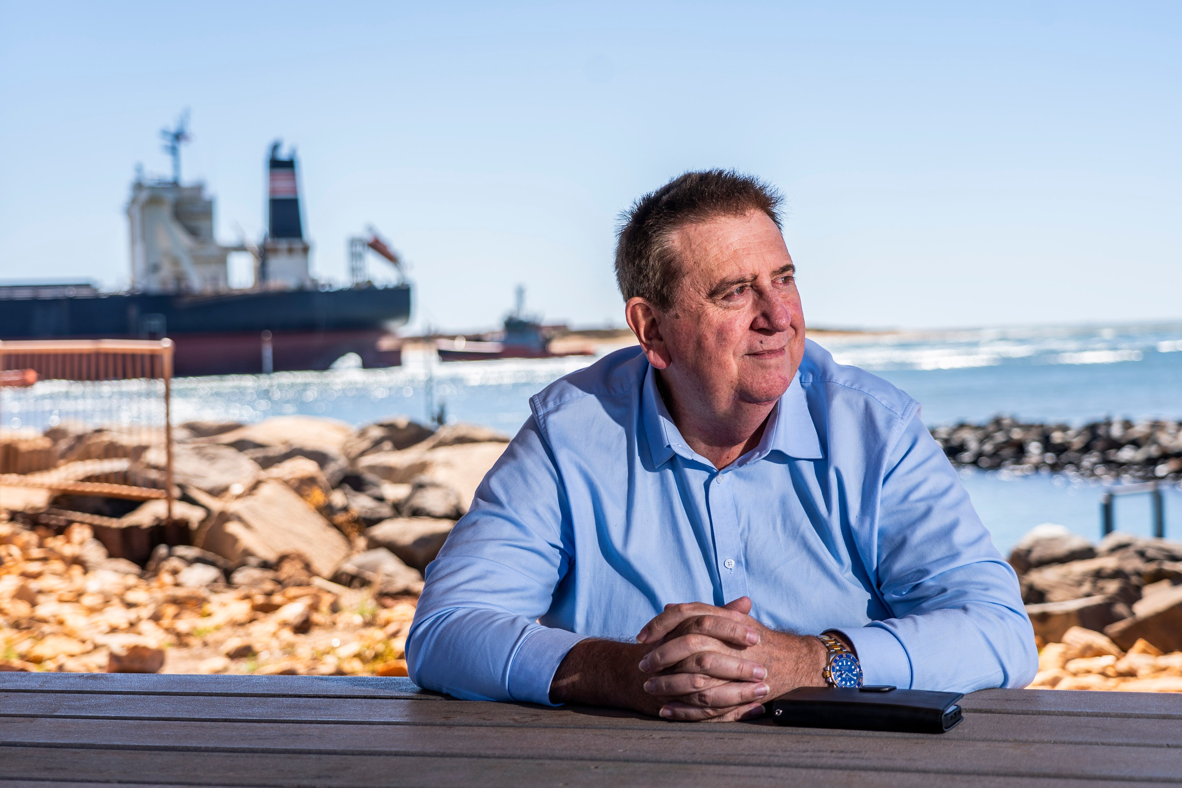 A middle-aged, dark-haired man sits in front of a rocky foreshore near where a bulk carrier is moored.