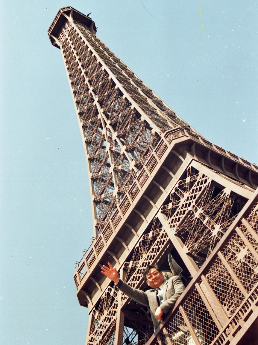 Against a clear blue sky, you view a miniature version of the Eiffel Tower, with a man at its base waving to the camera.