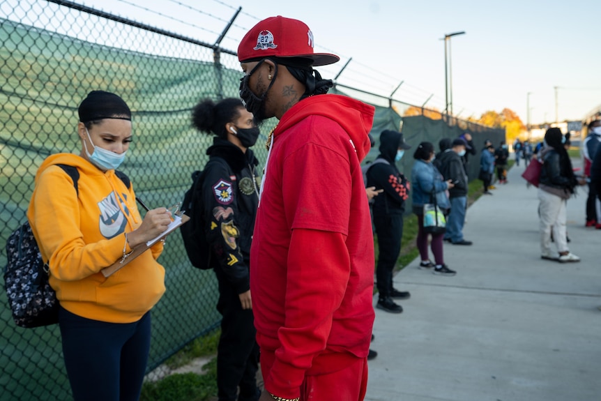 A man dressed in a red jumper approaches people in a line with a form.