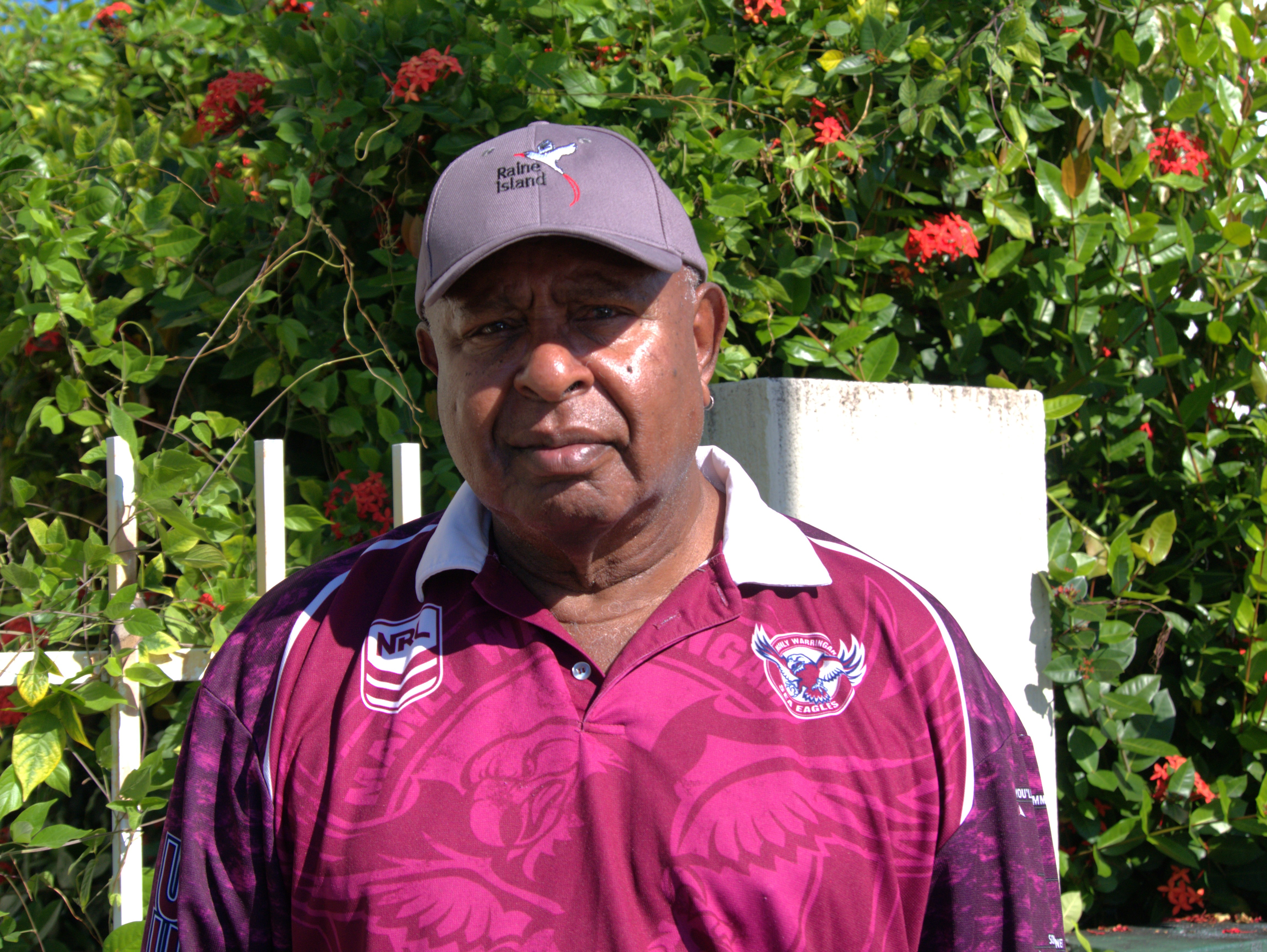 Falen with a serious face wearing a hat that says "Raine Island", a fuchsia pink and purple polo neck shirt with patterns.