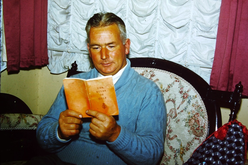 A middle aged man sitting in a chair reading a book called "Rules of Golf"