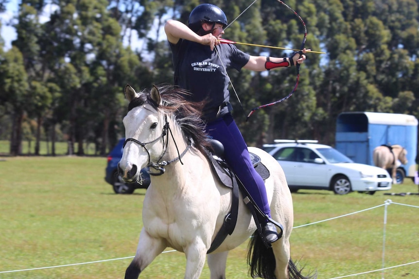 A woman on a white horse pointing a bow and arrow