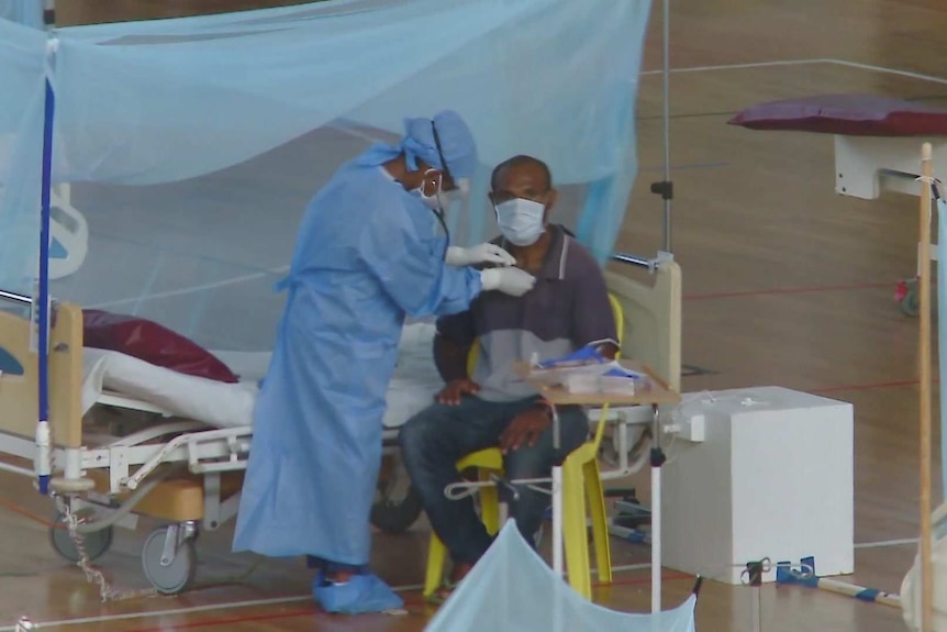 A seated man in a mask is treated by a person in full PPE next to a bed in an indoor sports stadium.