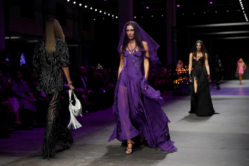 A model on a runway walks in a purple floor-length gown and a matching veil over her face.