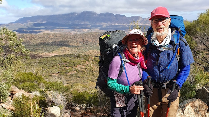 A man and a woman in hiking gear, including back packs, with bush and mountains in the background
