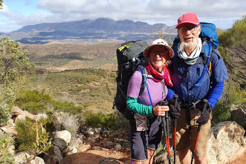 A man and a woman in hiking gear, including back packs, with bush and mountains in the background