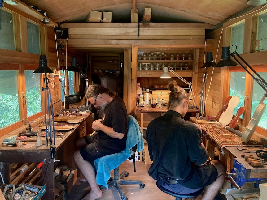 Two men sit back-to-back making violins in a mobile workshop