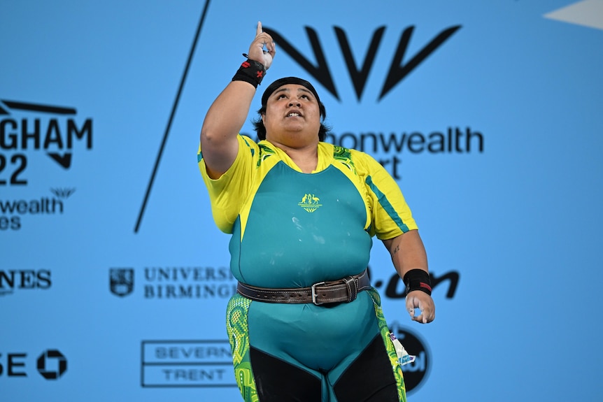 An Australian weightlifter stands on stage and points her finger to the sky after clinching a Commonwealth Games medal.