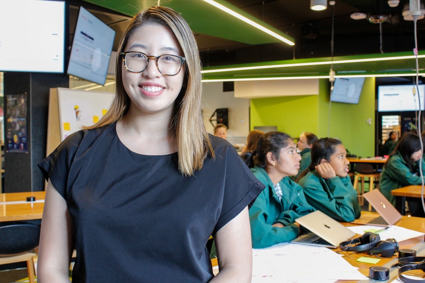 An asian woman with glasses stares smiling at the camera, students sit behind looking right of frame