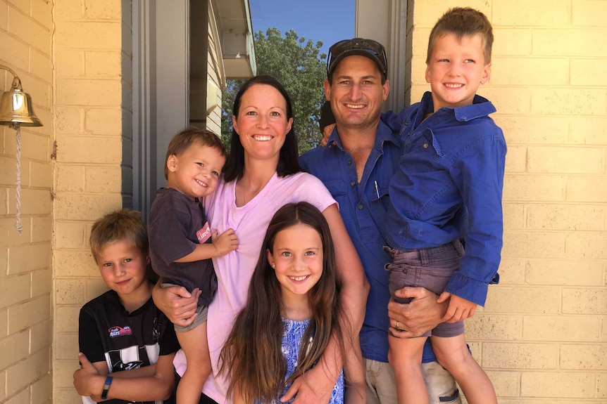 A family of a man, woman, three young boys, and a young girl, stand smiling at a camera in front of a yellow-brick home.