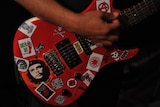 A hand strums a red electric guitar that is covered in stickers