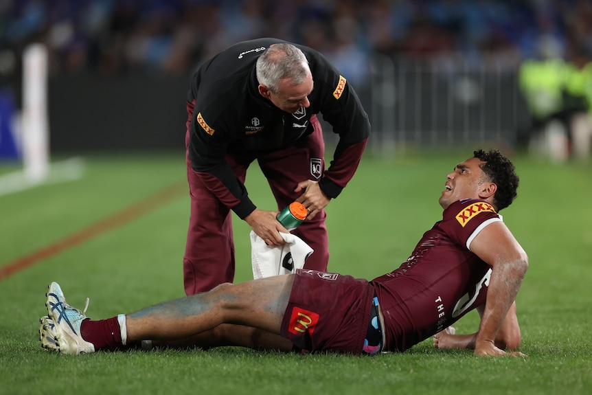 A man in maroon lies on the ground and looks at a trainer while in obvious pain.